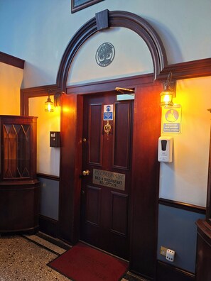A hotel entrance with a wooden door, a welcome mat, and hand sanitizer dispenser. - Pillars House (Edinburgh)