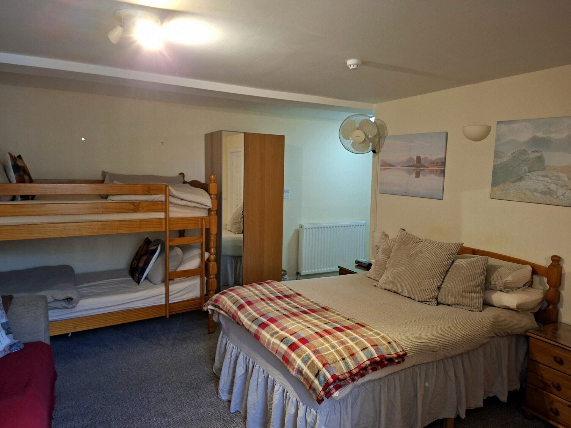 A bedroom with a bunk bed, a single bed, a bedside table, a lamp, and a wall-mounted fan.