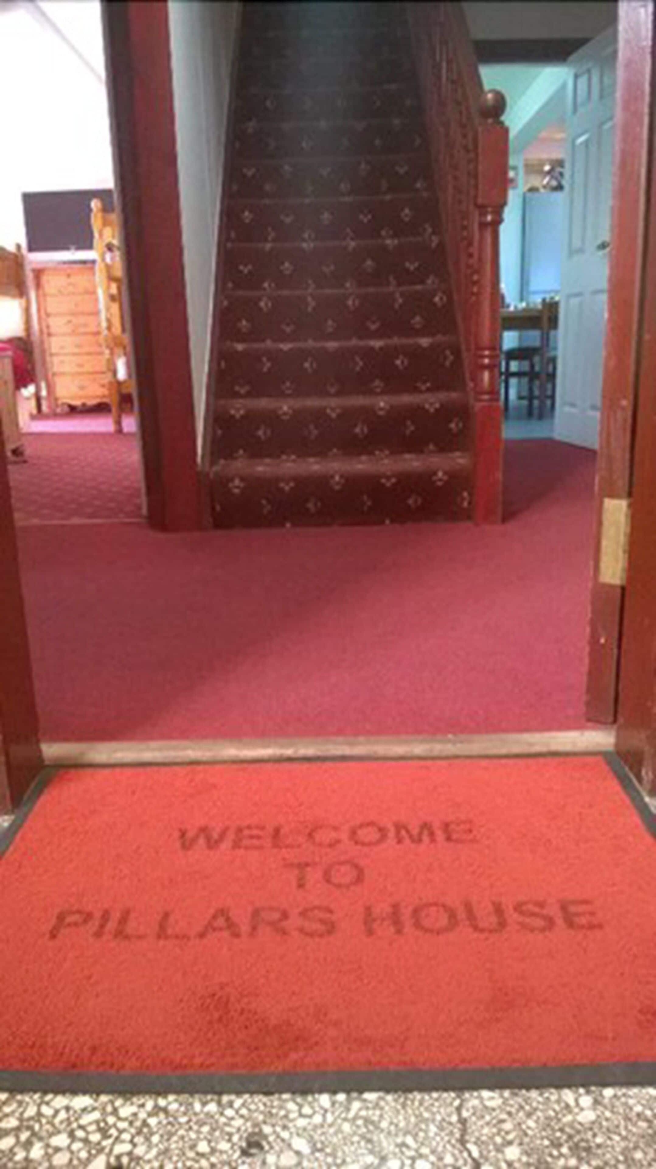 Ein roter Teppich führt zu einer Treppe, vor der ein Willkommensteppich mit der Aufschrift 'WELCOME TO PILLARS HOUSE' liegt.