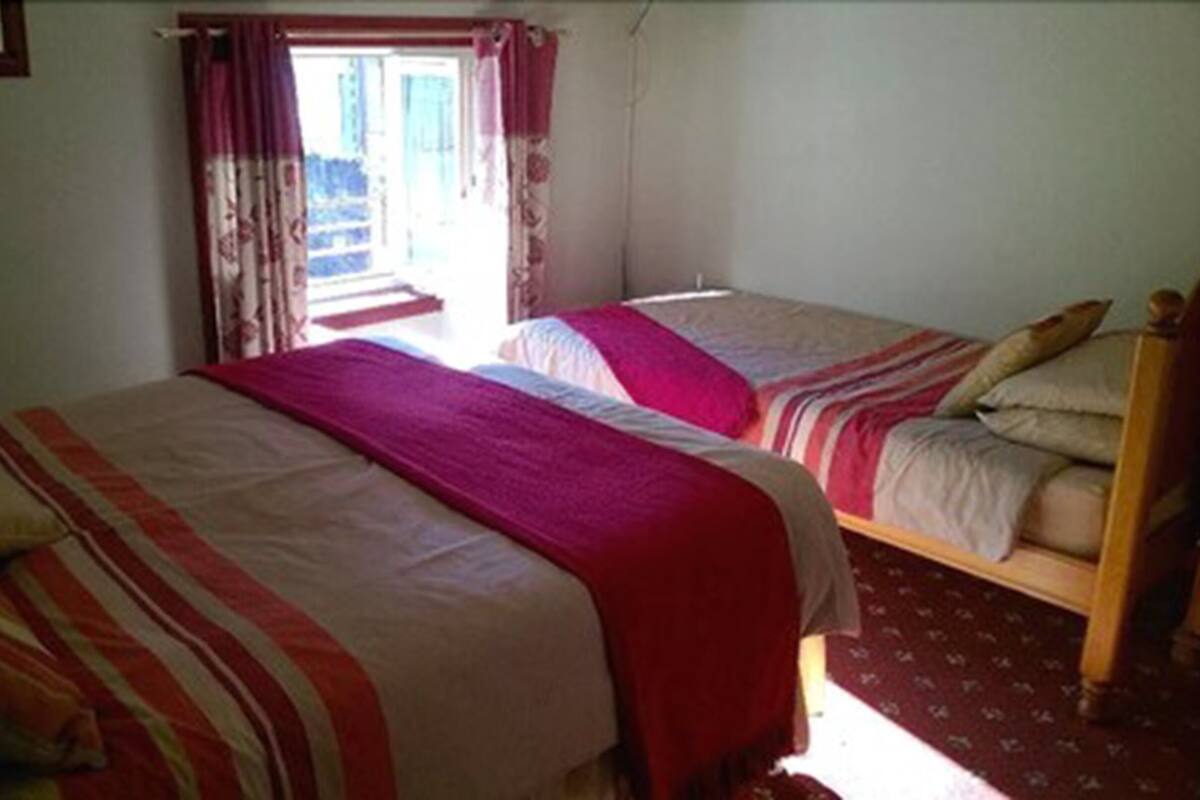 Quarto com duas camas, cada uma com roupa de cama e almofadas listradas. Há uma janela com cortinas, e uma porta que dá para o exterior.