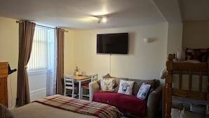 A cozy room with a bed, a sofa, a wooden chair, a small table, and a TV mounted on the wall.