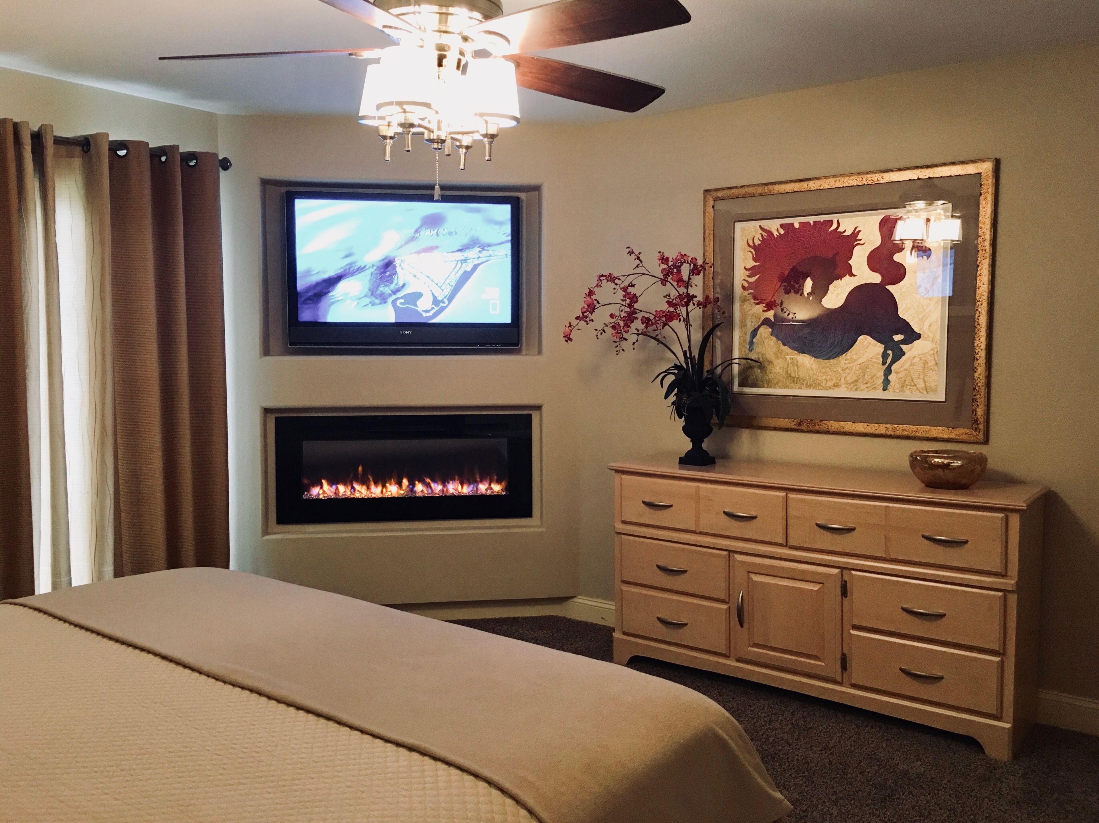A hotel room with a bed, a television mounted above a fireplace, a dresser, and a framed artwork on the wall.