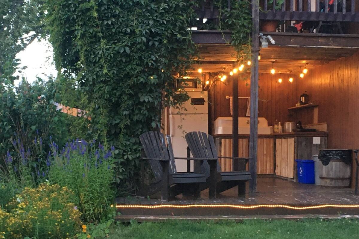 An outdoor seating area with wooden chairs and a table, surrounded by greenery and string lights.