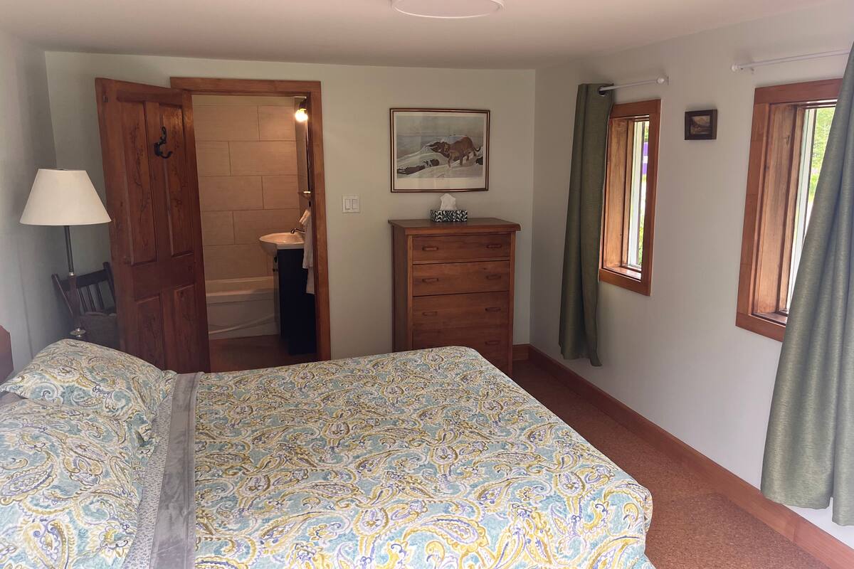 A bedroom with a bed, a wooden dresser, a nightstand with a lamp, a bathroom visible through an open door, and a window with curtains.