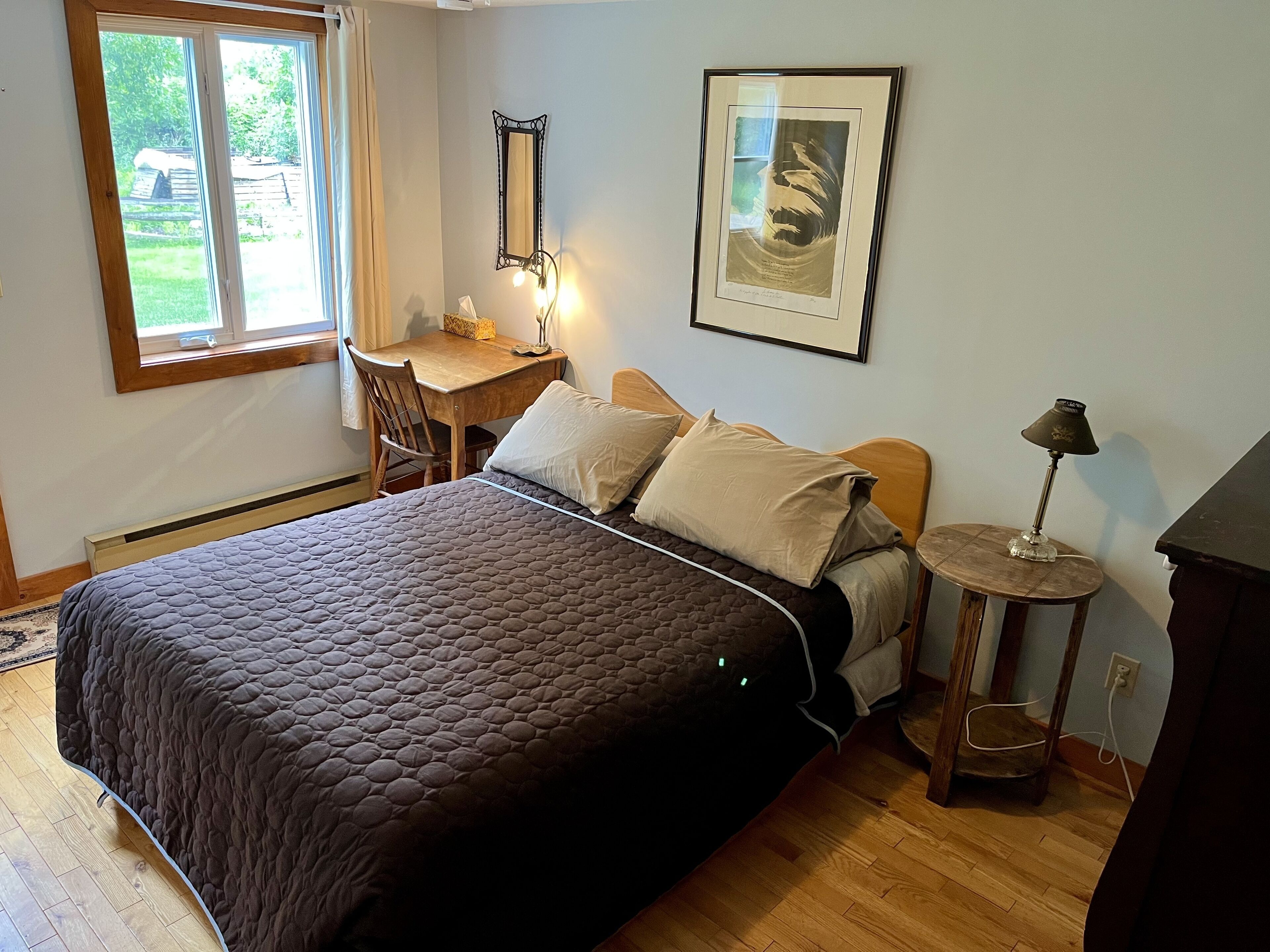 A bedroom with a large bed, a desk, a chair, a small table with a lamp, and a framed picture on the wall.