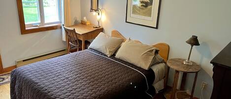A bedroom with a large bed, a desk, a chair, a small table with a lamp, and a framed picture on the wall.