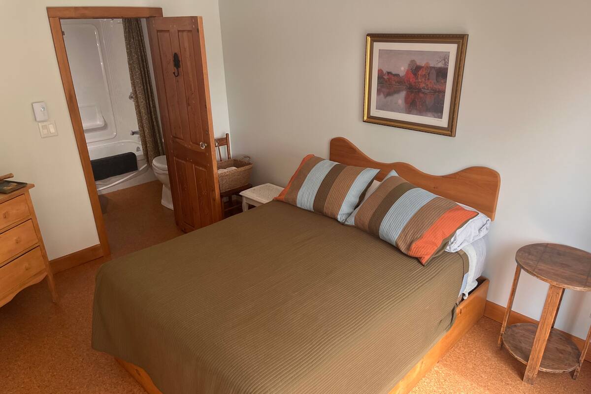 A bedroom with a large bed, a wooden headboard, a small bedside table, a wooden nightstand, a framed picture on the wall, and a door leading to a bathroom.