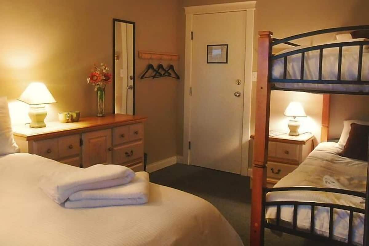 Habitación de hotel con una cama individual, una mesita de noche con lámpara, un aparador con espejo y una puerta con perchero.