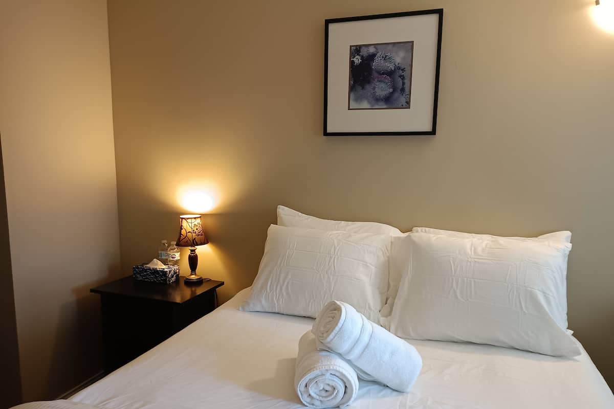 Una cama bien hecha con sábanas y almohadas blancas, una toalla enrollada sobre la cama, una mesita de noche con una lámpara y una caja de pañuelos, y un cuadro en la pared.