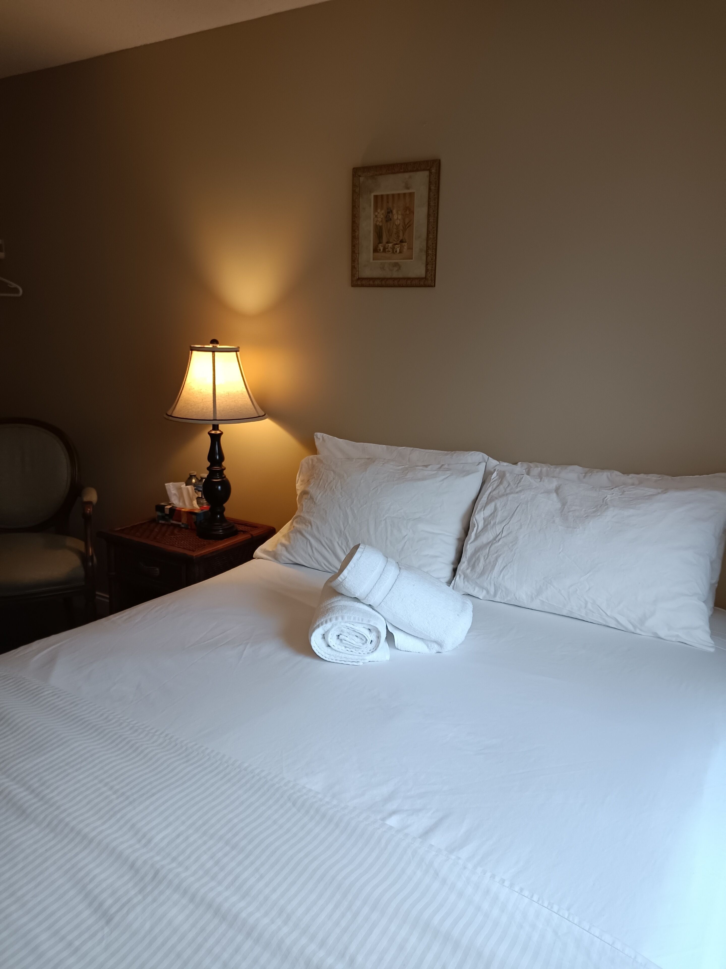 A neatly made bed with white linens, a rolled towel, a bedside lamp, and a framed picture on the wall.