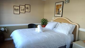 A neatly made bed with white linens, a headboard, and a bedside table with a lamp. There are three framed pictures on the wall and a potted plant beside the bed.