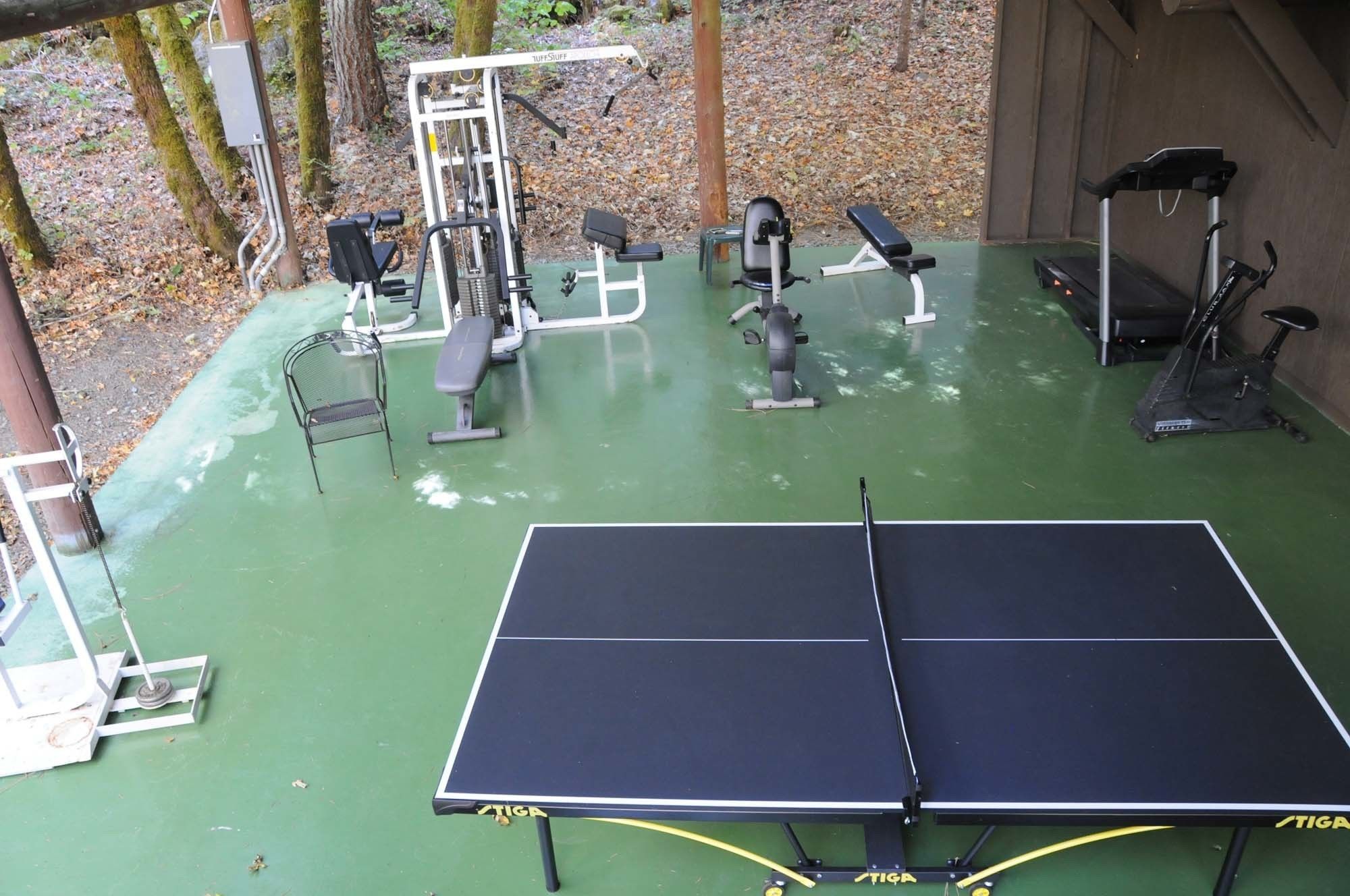 Un espacio exterior techado para gimnasia, con mesa de ping pong, equipo de ejercicios y bancos.