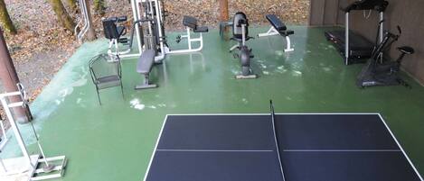 Un espacio exterior techado para gimnasia, con mesa de ping pong, equipo de ejercicios y bancos.