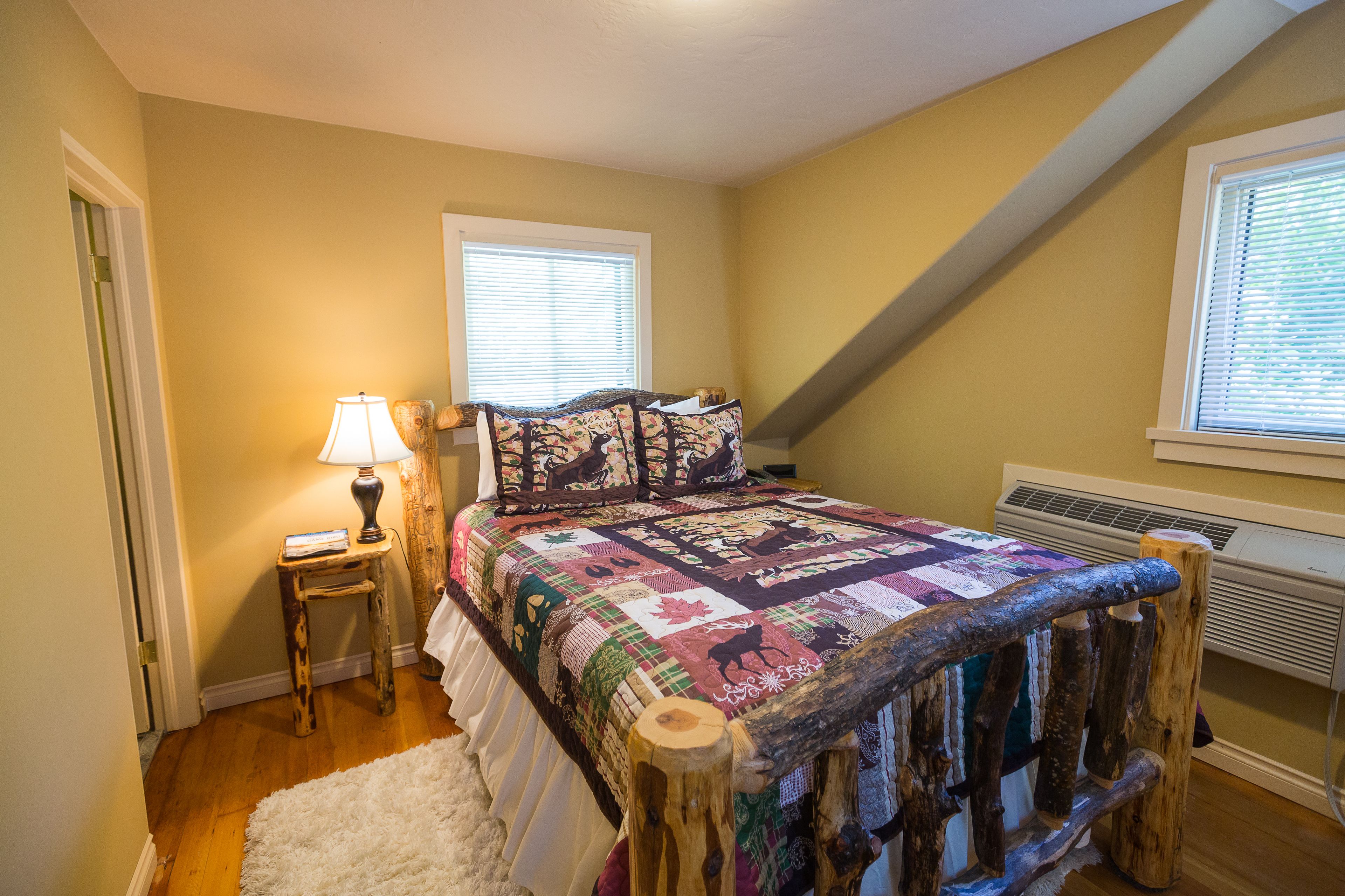 Une chambre à coucher avec un lit en bois, une courtepointe, une table de chevet avec une lampe et une fenêtre avec des stores.