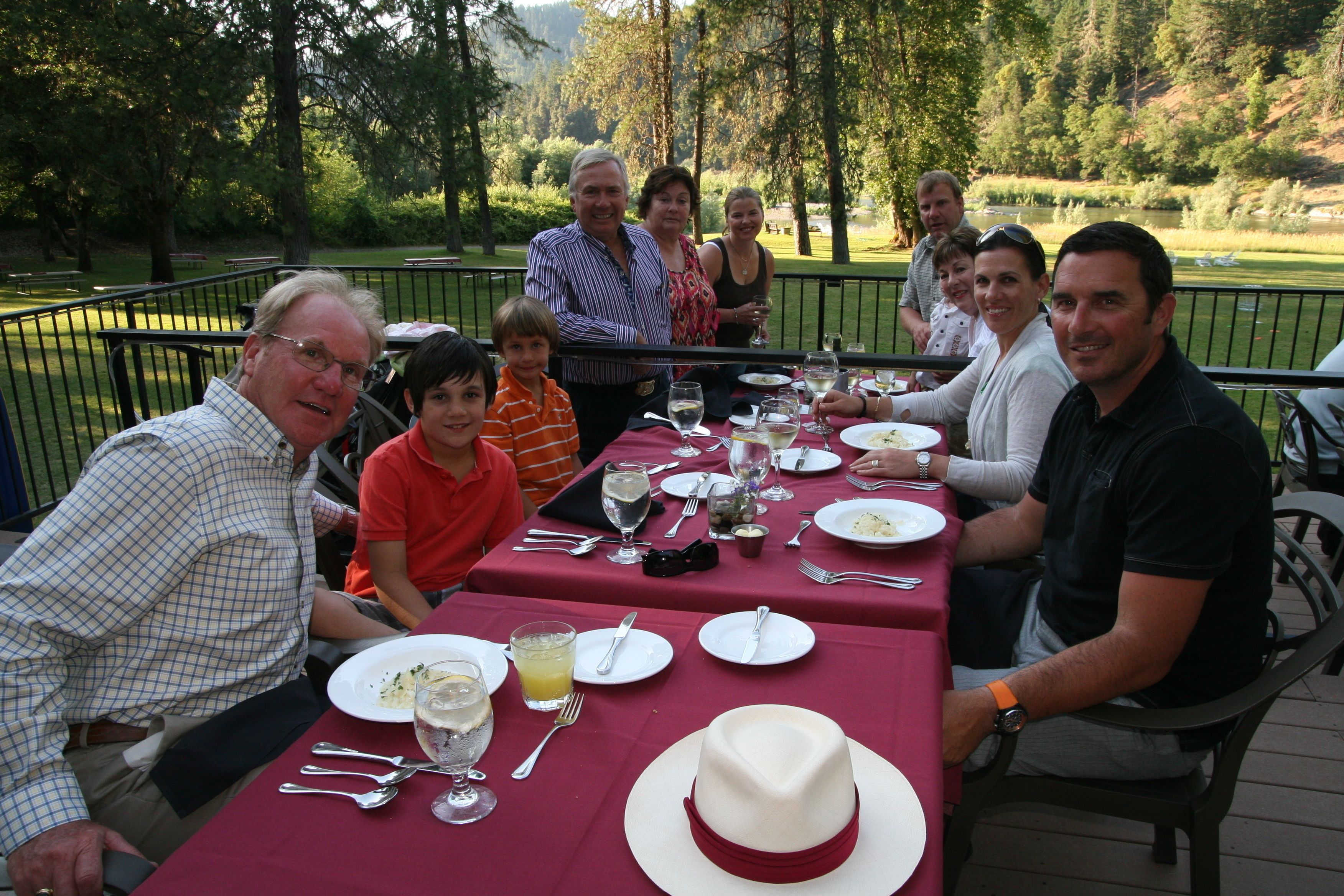 Un grupo de personas cenando al aire libre en una mesa con mantel rojo, rodeado por un parque pintoresco y vistas a un lago.