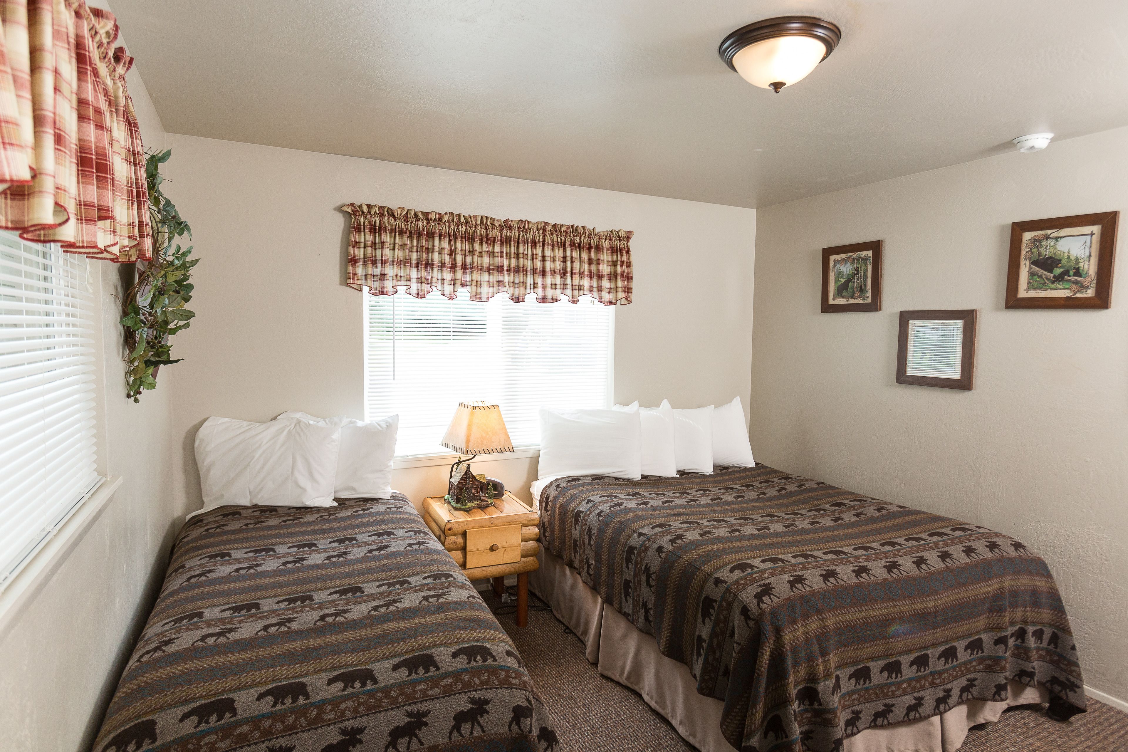 A room with two beds, a window with blinds, a lamp on a nightstand, and wall-mounted pictures.