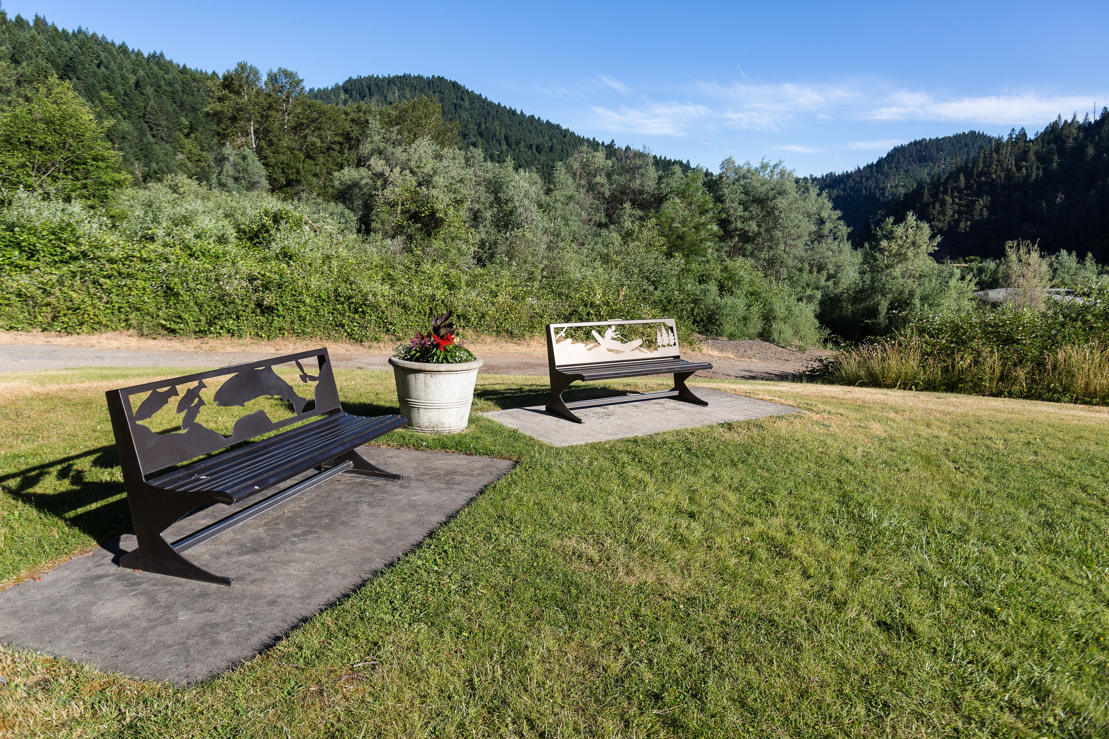 Two benches in a park with a grassy area, a concrete path, and a backdrop of dense forest and hills.