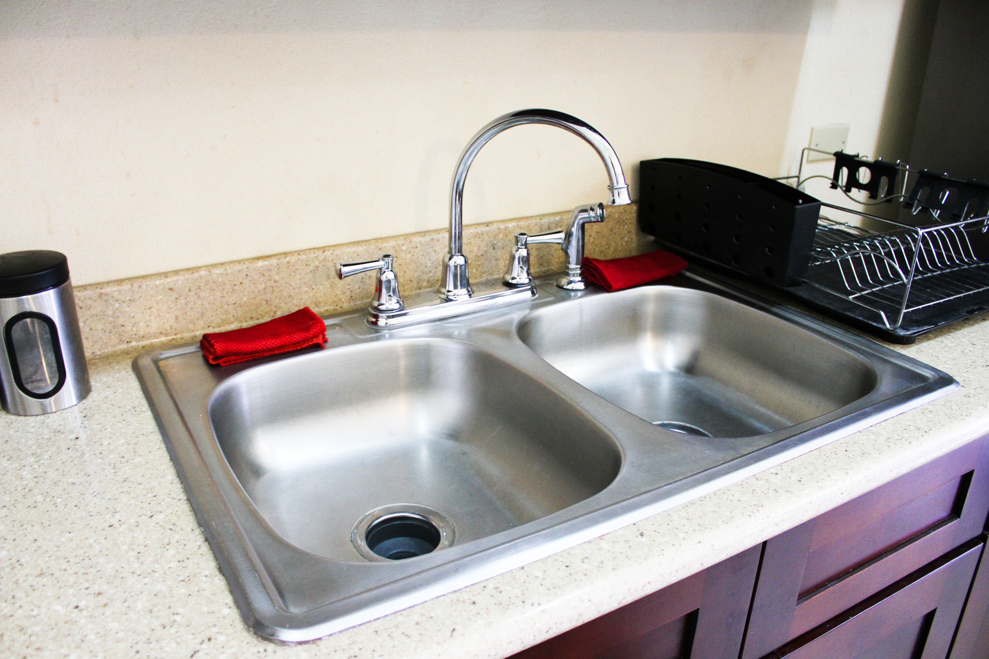 A stainless steel kitchen sink with a double faucet, a red cloth, and a coffee maker on the counter.