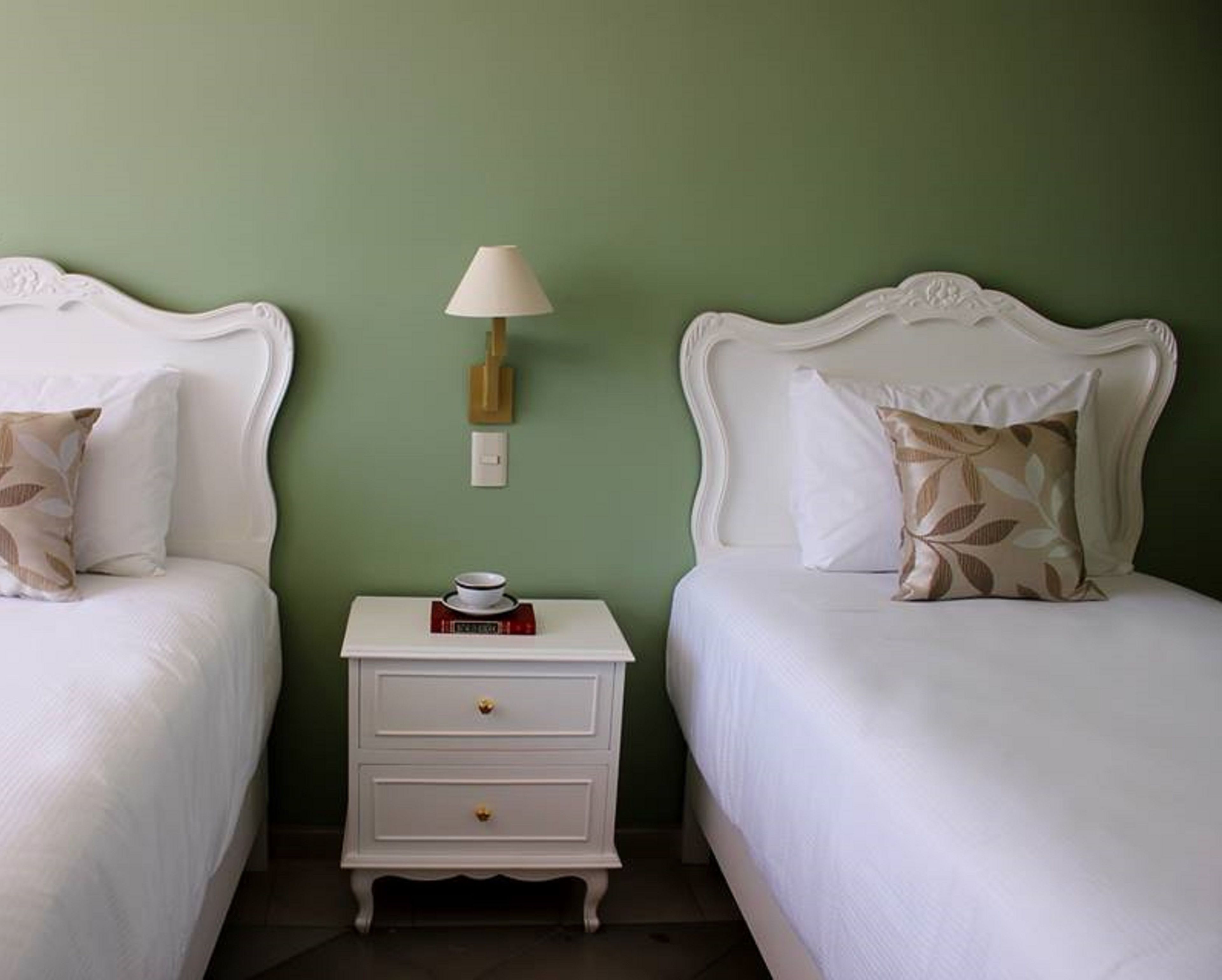 A room with two white beds, a white nightstand with drawers, a wall-mounted lamp, and a small table with a cup and book.