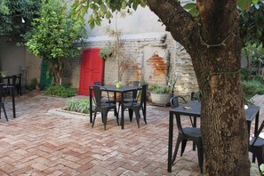Un patio al aire libre con suelo de ladrillo, mesas y sillas de metal negro, una puerta roja y plantas en macetas.