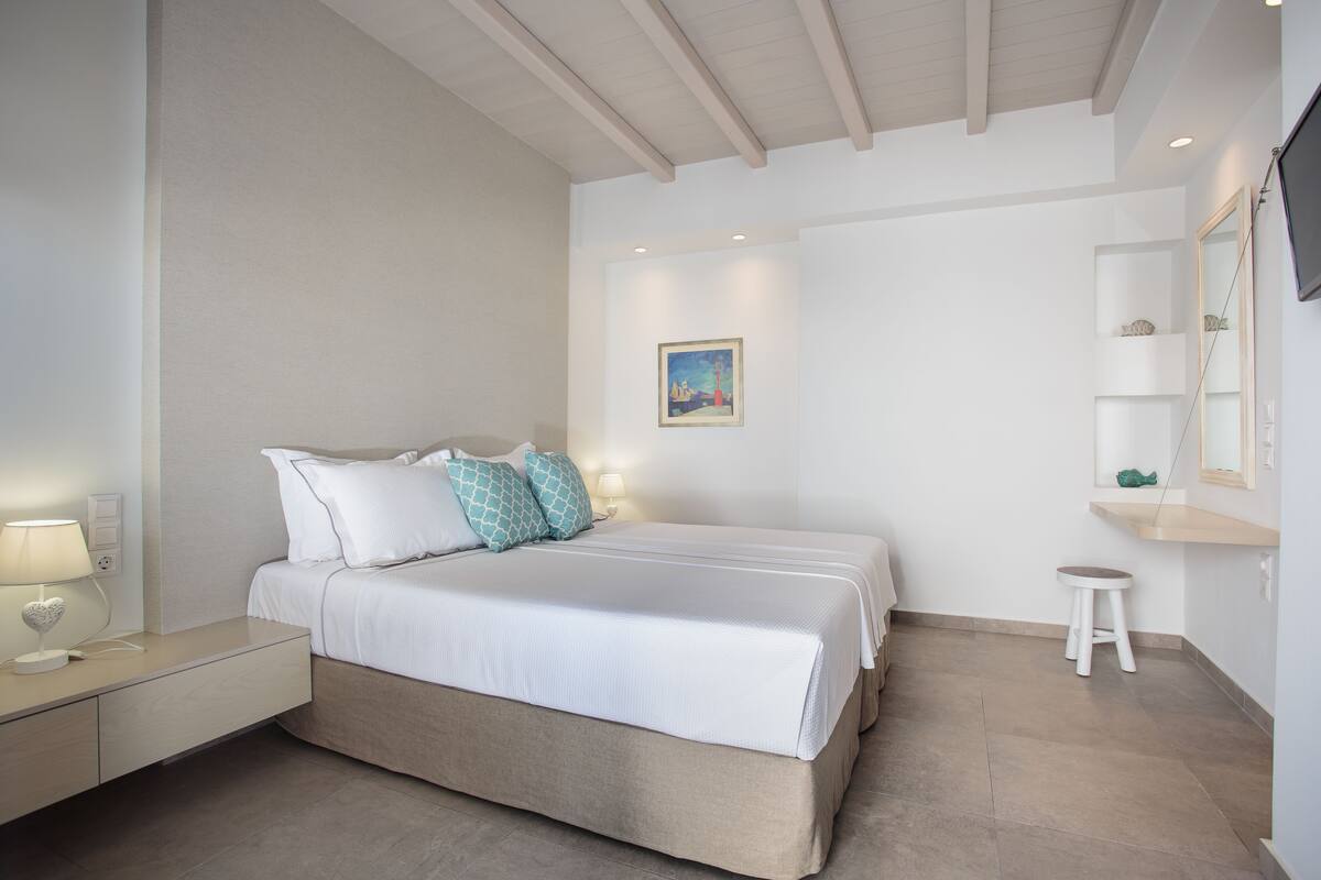 Une chambre à coucher comprenant un lit, des tables de chevet, une commode, un petit tabouret, un tableau au mur et une étagère avec des objets décoratifs.