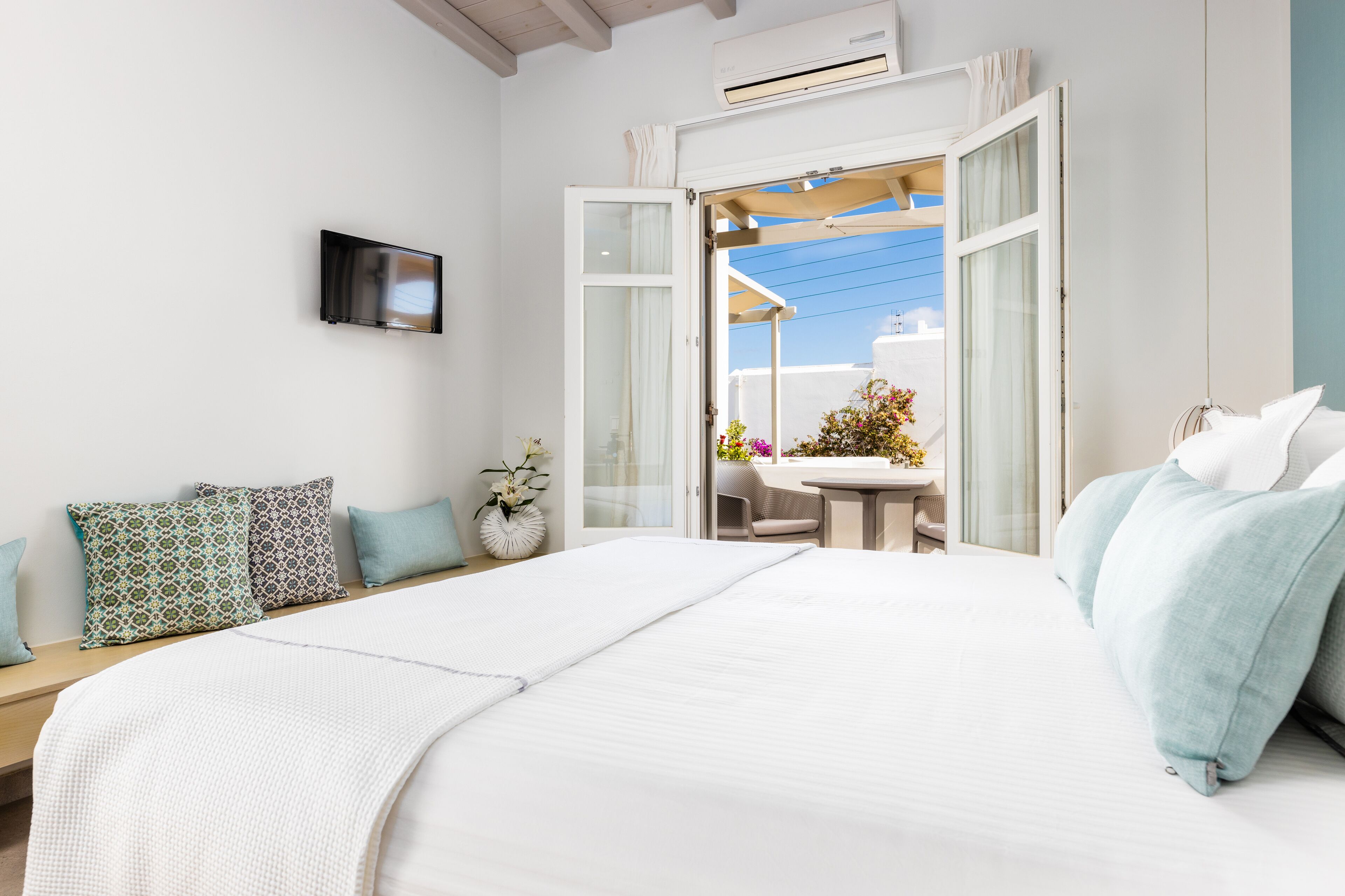 A bedroom with a large bed, a bedside table, a TV mounted on the wall, and an open door leading to a balcony with a table and chairs.