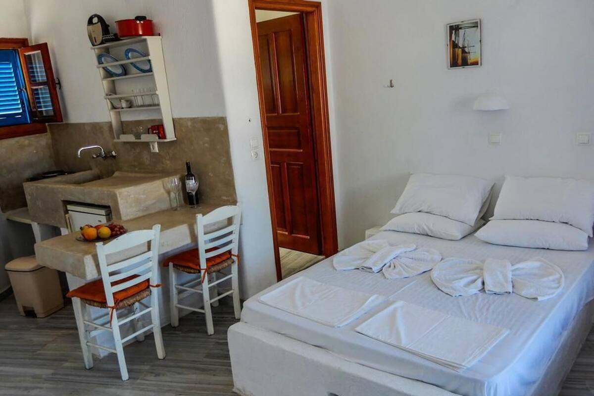 A bedroom with a bed, a small desk, and a chair. There is a wooden door and a shelf with kitchen items.