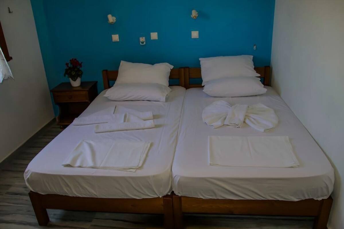 A room with two beds, each with white linens and pillows, a small bedside table with a plant, and a blue wall with light switches.