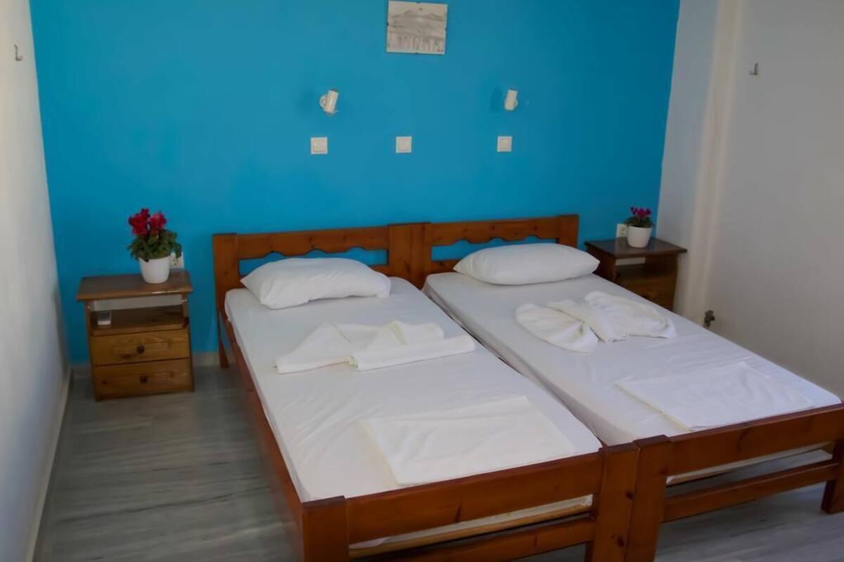 A room with two wooden beds, white bedding, a blue wall, and a small table with a potted plant.