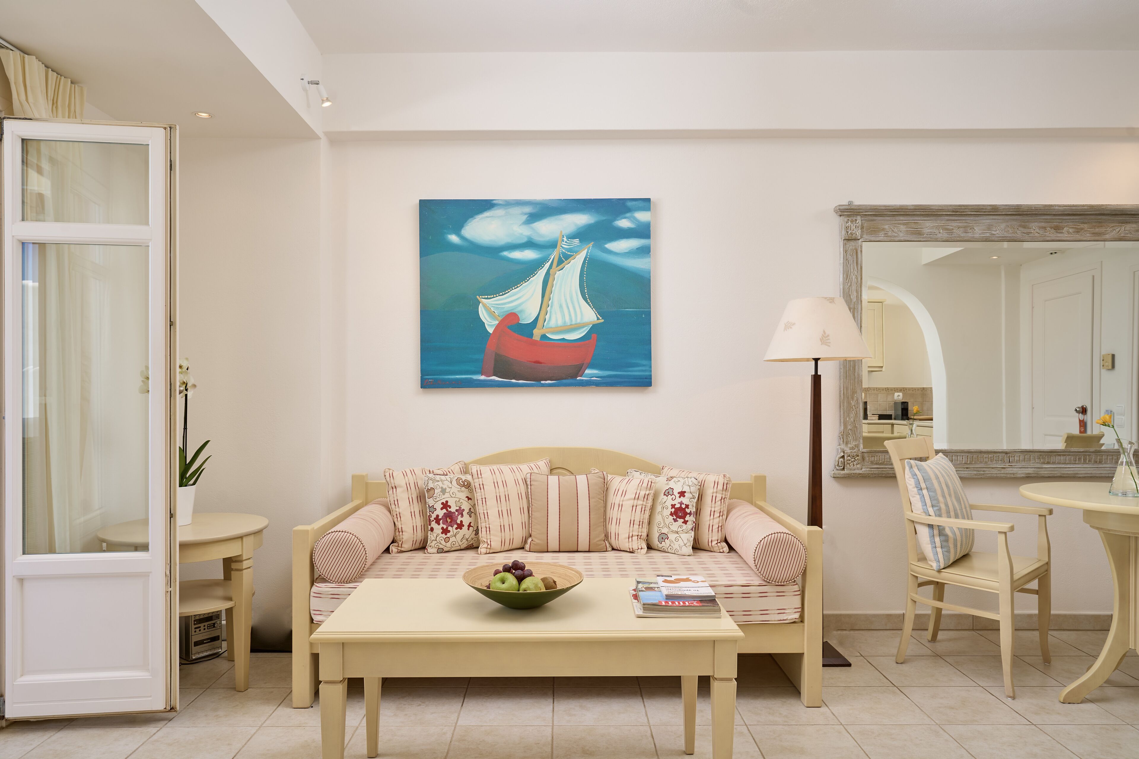 A living room with a sofa, a coffee table with a bowl of fruit, a painting of a ship on the wall, and a mirror above a door.