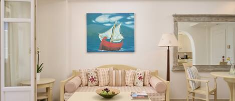 A living room with a sofa, a coffee table with a bowl of fruit, a painting of a ship on the wall, and a mirror above a door.