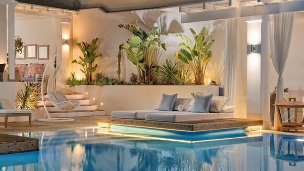 A modern indoor pool area with a lounge chair, a sofa with cushions, and a variety of plants. - Stelia Mare Boutique Hotel (Paros)