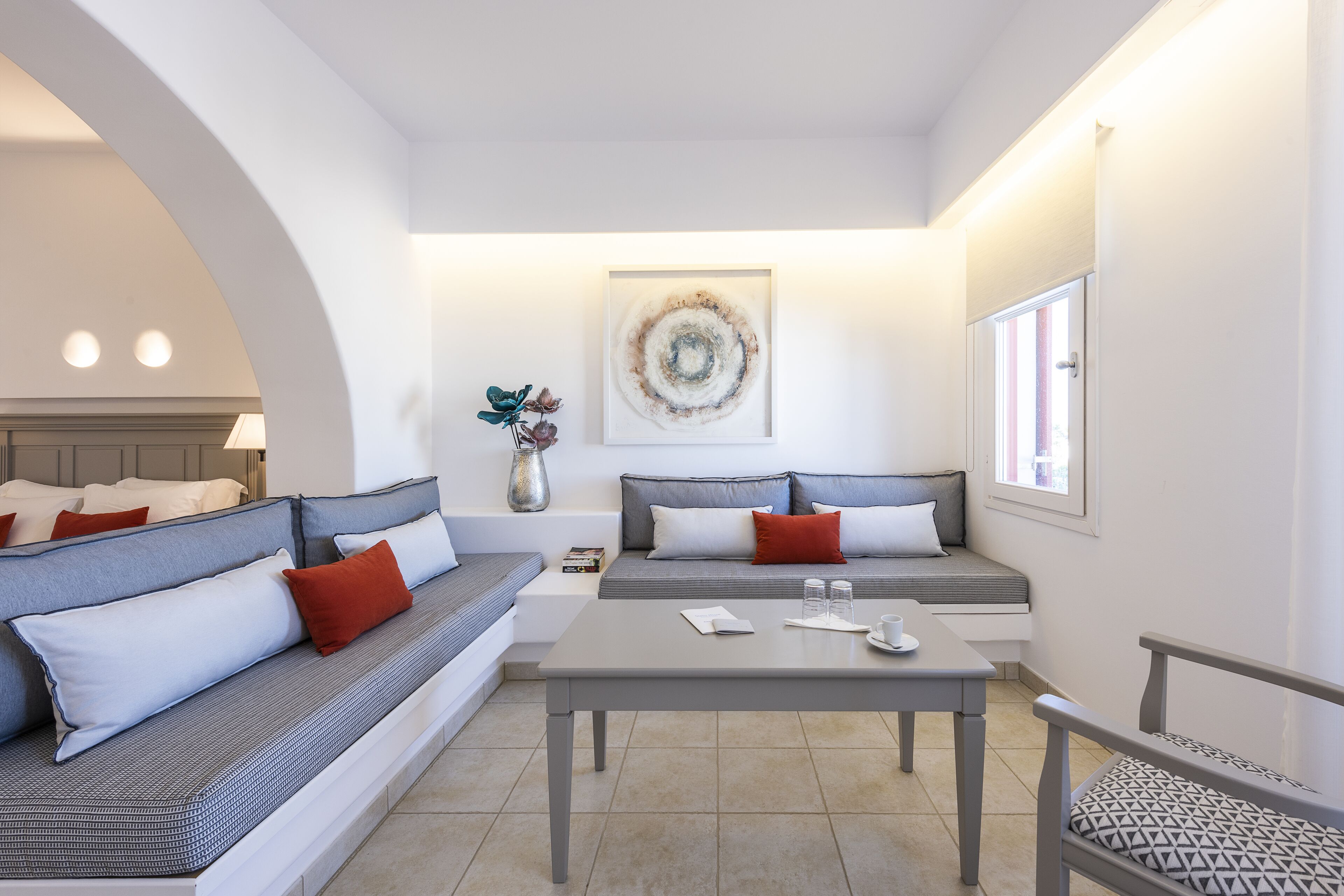 A modern living room with a sofa, a table, and a chair. There is a painting on the wall and a window with curtains.