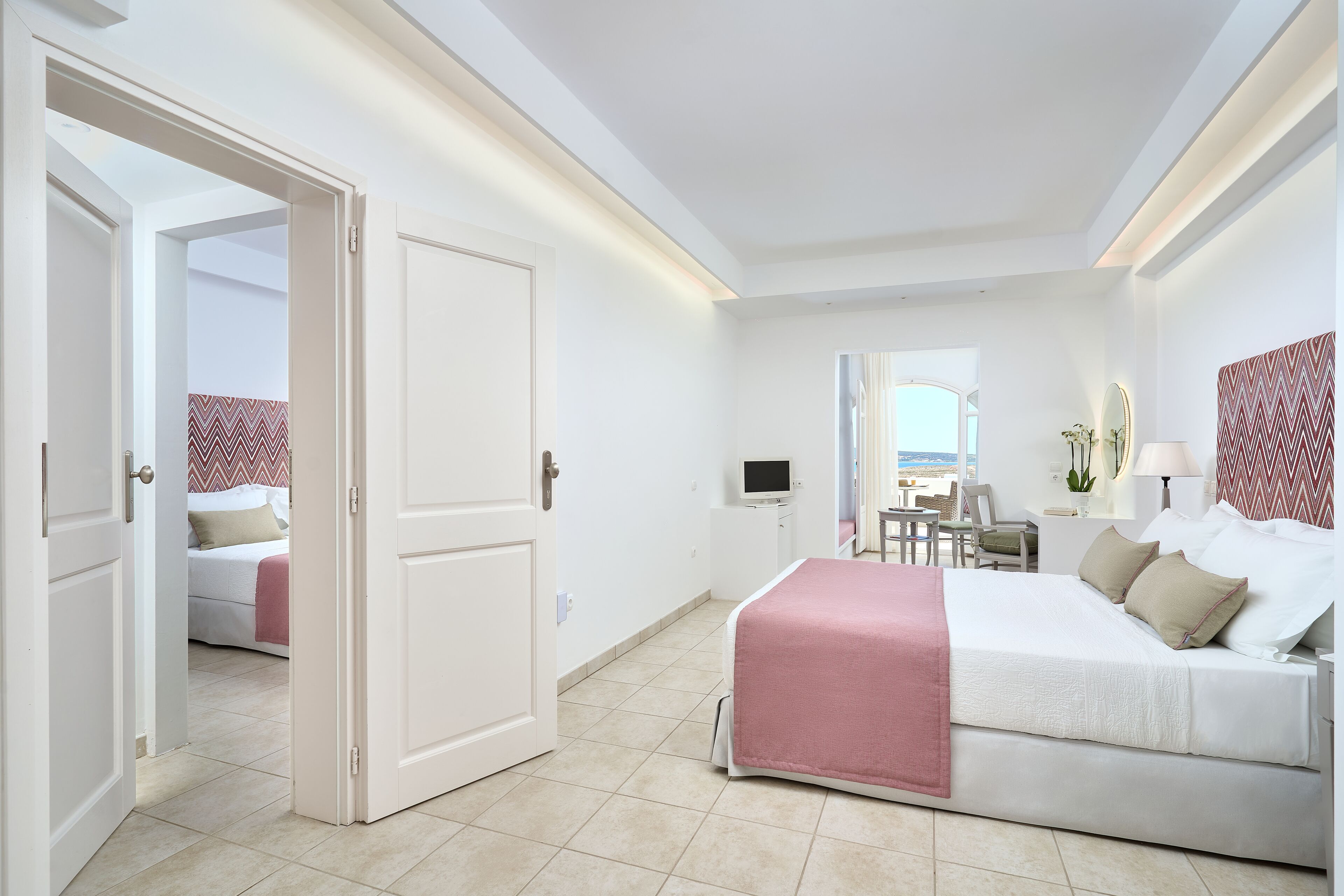 A hotel room with a bed, a desk, a chair, a TV, and a view of the sea.