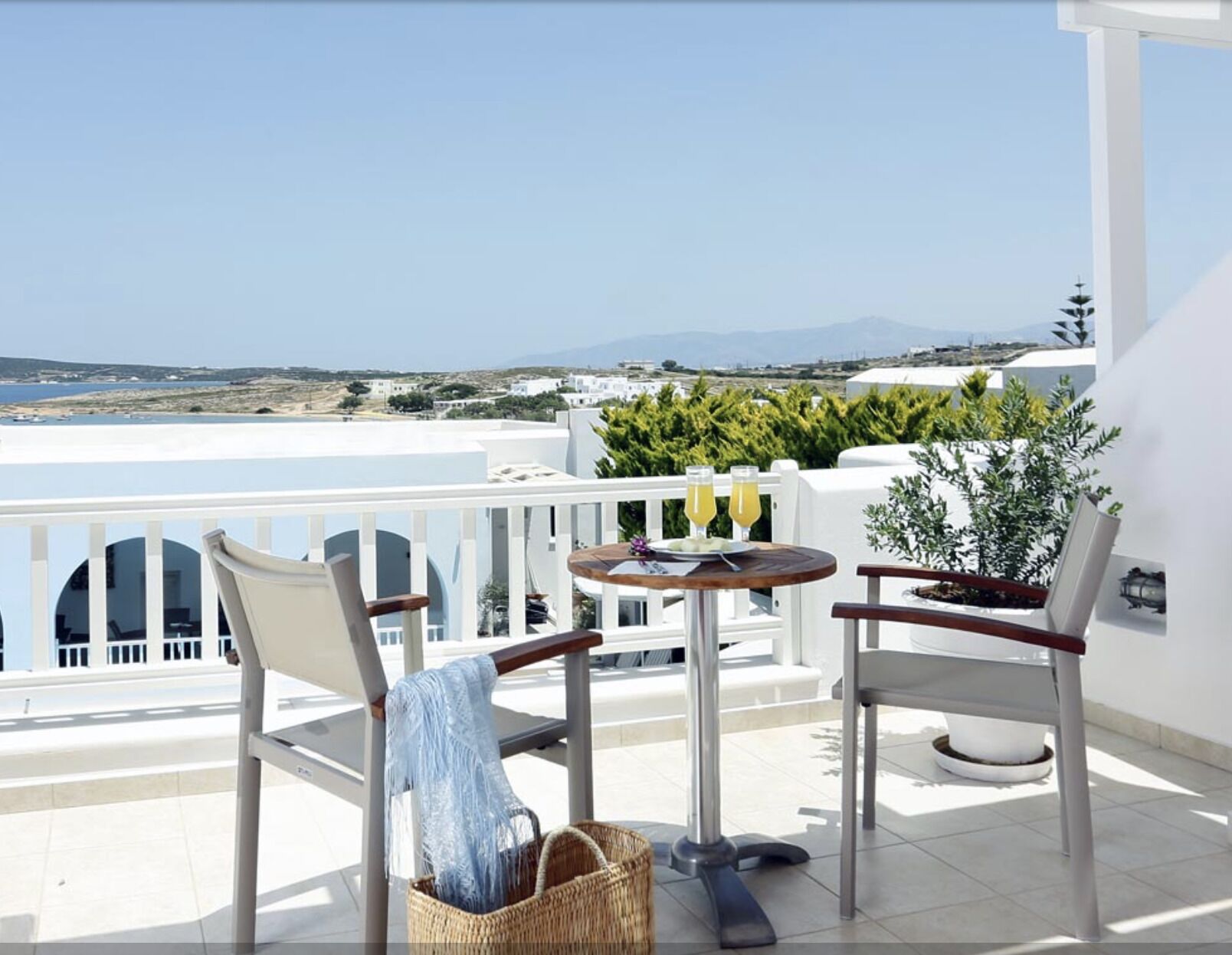A balcony with two chairs, a small table, and a basket. The balcony overlooks a coastal landscape with buildings and a clear sky.