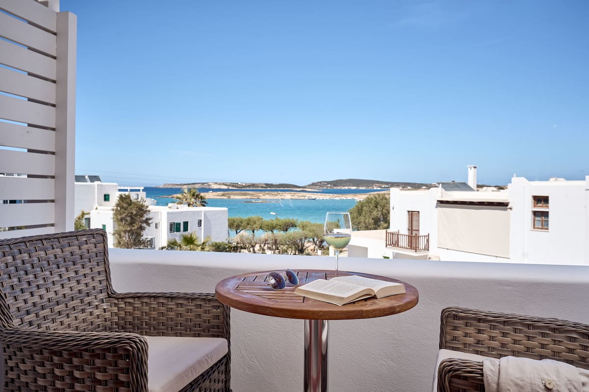 A balcony with wicker chairs, a small round table with a book and sunglasses, and a glass of wine. The view includes a coastal landscape with buildings and the sea.