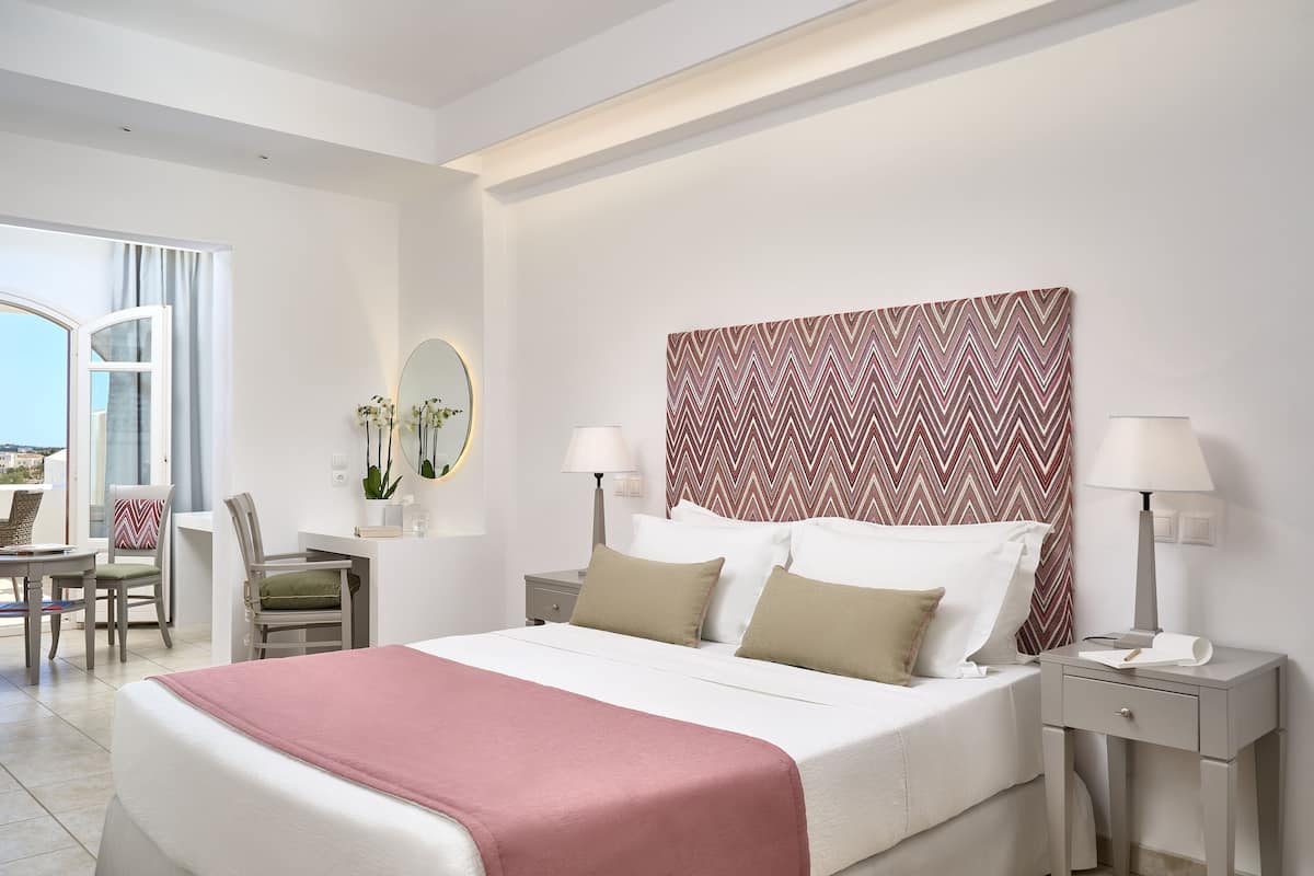 A hotel room with a bed, bedside tables, a desk, chairs, a window, and a patterned wall.