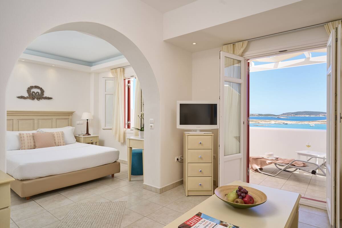 A hotel room with a bed, a TV, a desk with a bowl of fruit, and a balcony with a view of the sea.