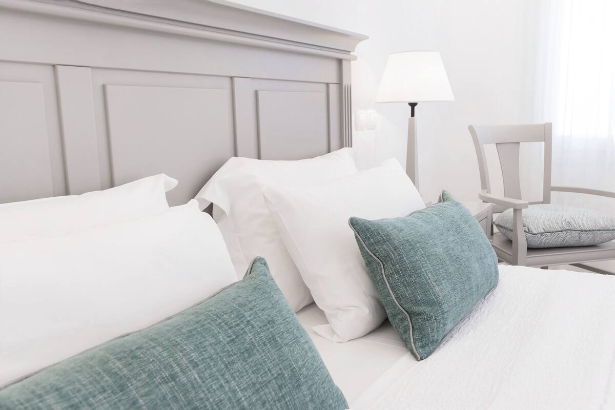 A neatly made bed with white and teal pillows, a white headboard, and a bedside table with a lamp and a chair.