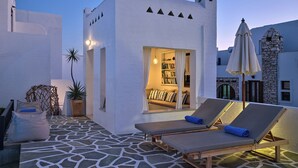Un edificio bianco con un balcone, una zona salotto con cuscini e una libreria visibile attraverso una finestra aperta.