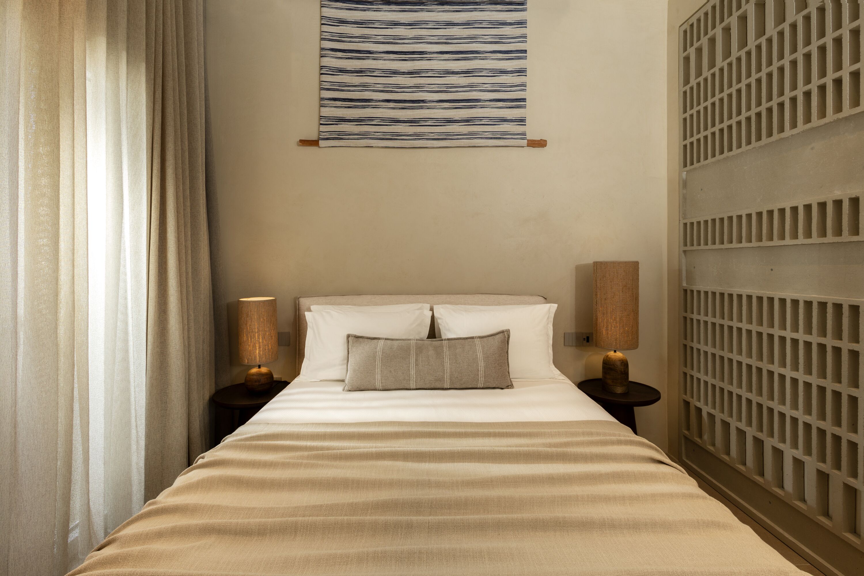 A hotel room with a bed, two bedside lamps, a headboard, a wall-mounted light fixture, and a window with curtains.