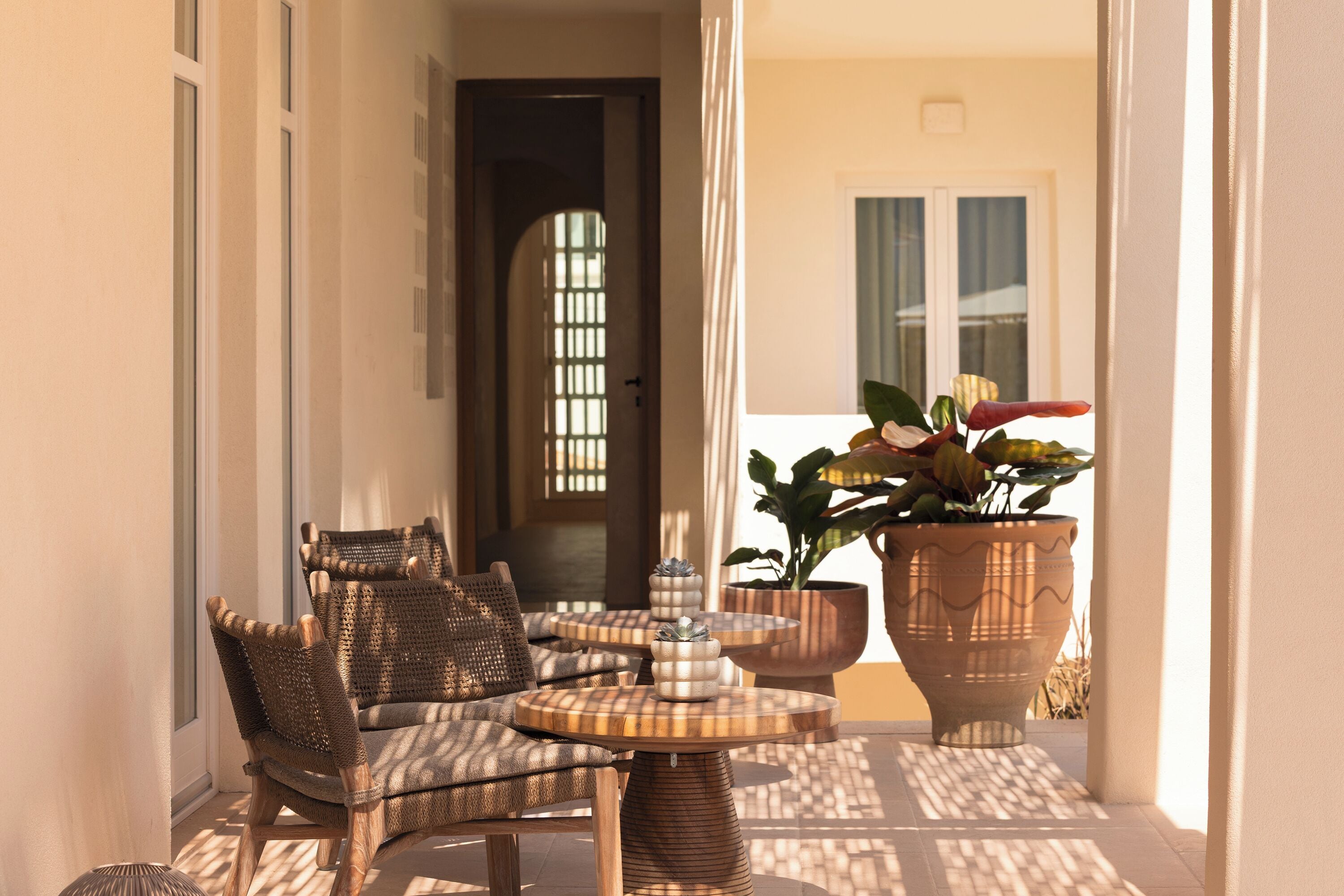 A sunlit outdoor seating area with wicker furniture and a round table, a potted plant, and a door leading to the interior.