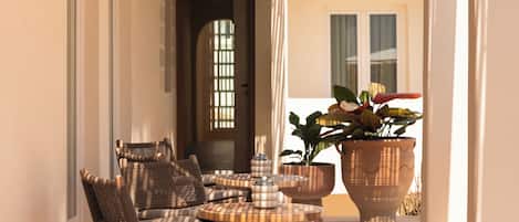 A sunlit outdoor seating area with wicker furniture and a round table, a potted plant, and a door leading to the interior.