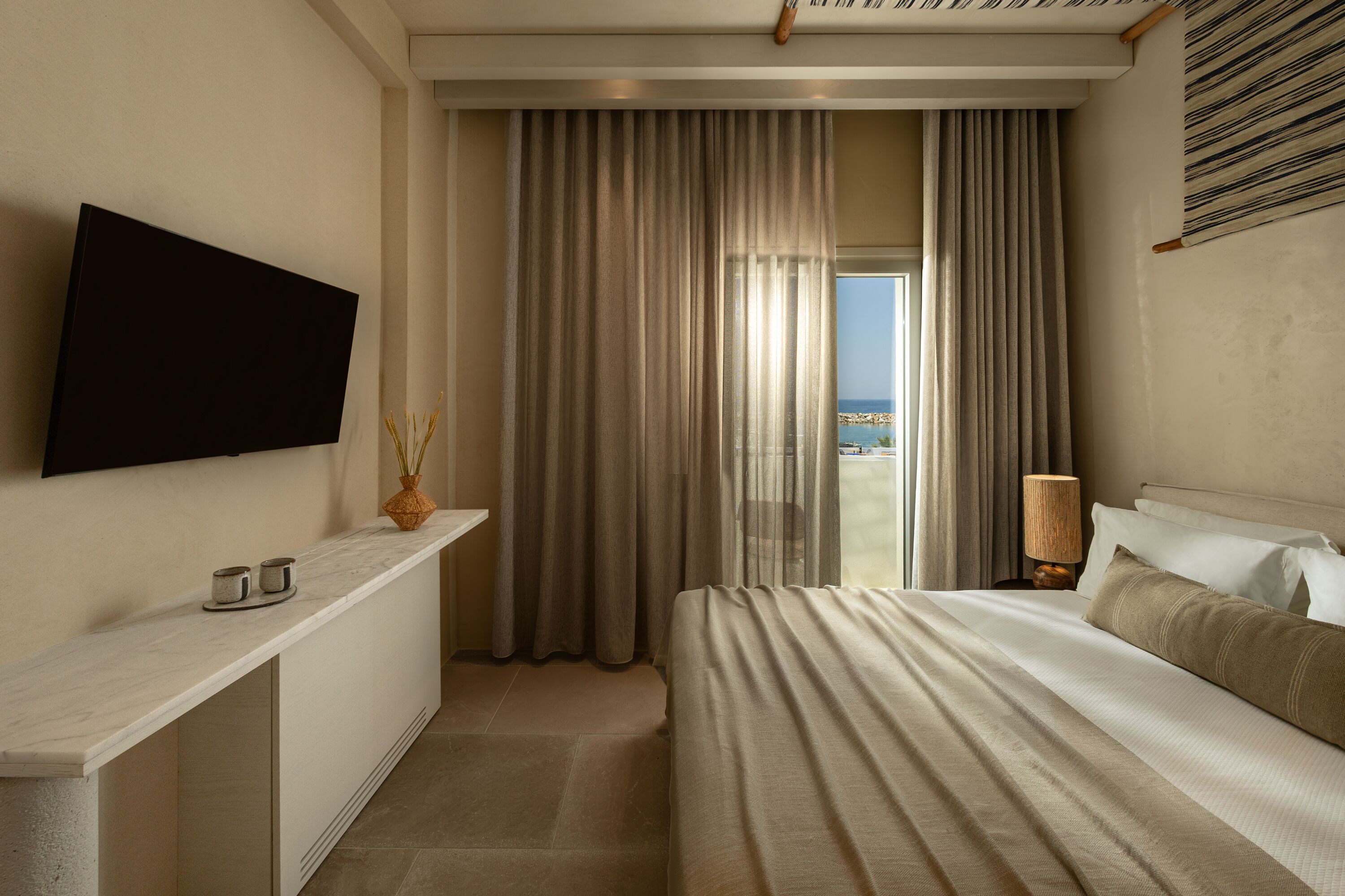 A hotel room with a bed, a TV mounted on the wall, a bedside table with a vase, and a view of the sea through the window.