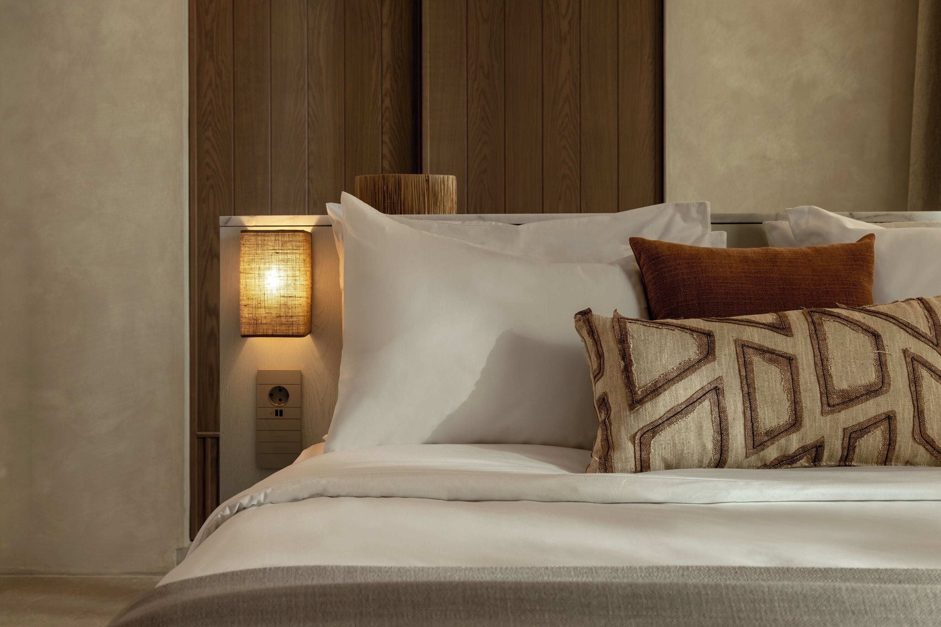 Een keurig opgemaakt bed met witte beddengoed en een kussen met een patroon, een wandlamp en een stopcontact op de muur.