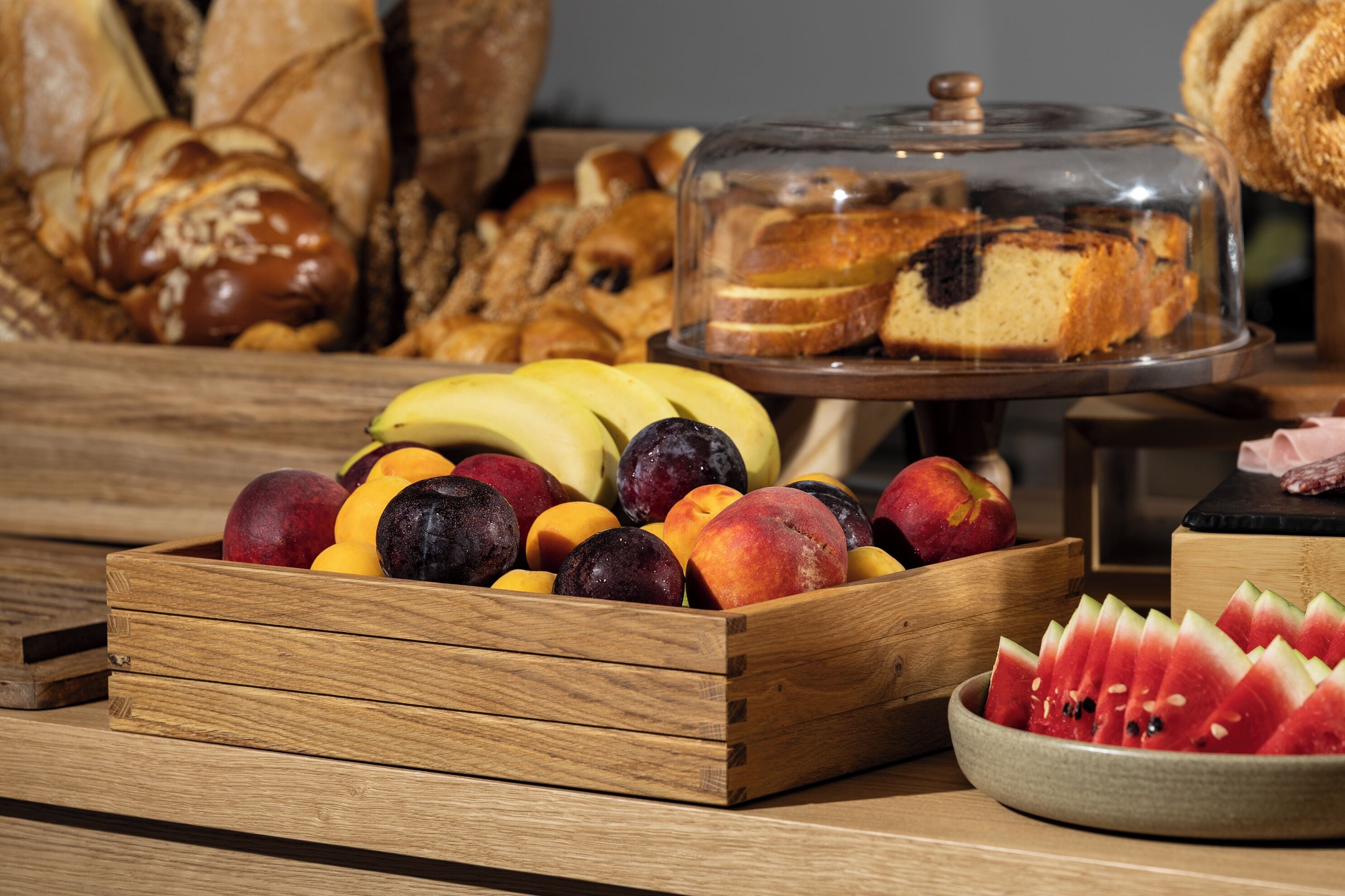 A wooden fruit crate filled with various fruits, a glass bowl of watermelon slices, and a glass display case with bread and a cake.
