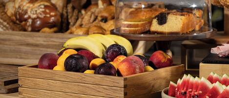 A wooden fruit crate filled with various fruits, a glass bowl of watermelon slices, and a glass display case with bread and a cake.