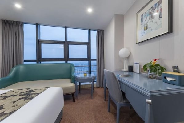 A hotel room with a bed, a desk, a chair, a small table, a potted plant, a framed picture, and a window with curtains.