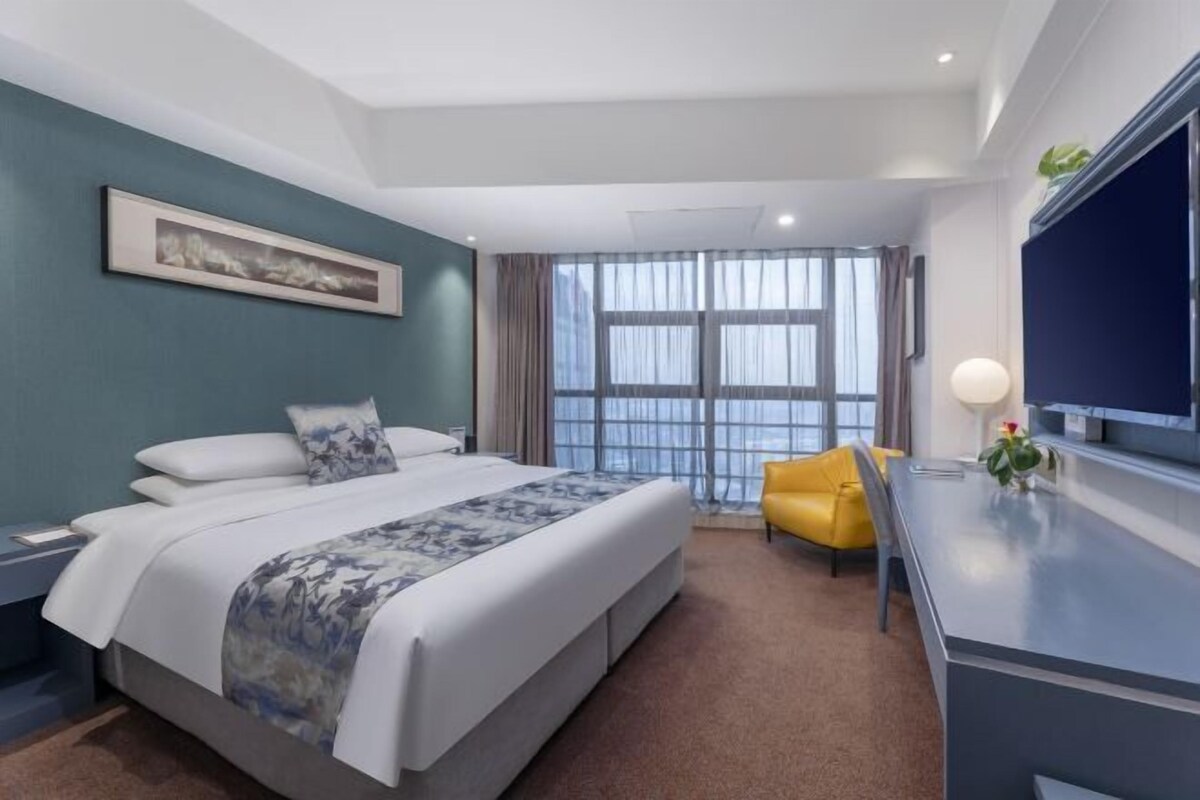 A modern hotel room with a large bed, a yellow armchair, a desk, and a large window with curtains.
