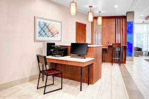 Ein moderner Arbeitsbereich mit Schreibtisch, Computer, Drucker und Stuhl. An der Wand hängt ein gerahmtes abstraktes Kunstwerk und es gibt einen Empfangsbereich mit einer Theke und Hocker.